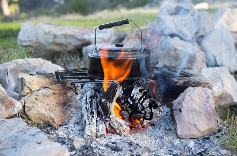 A campfire surrounded by rocks with flames coming out of logs and a campfire pot standing over the flames on a grill