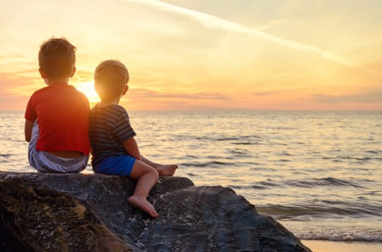 Two young boys sitting on a rock at the seashore watching the sun setting