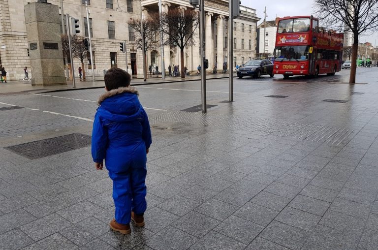 A picture of a boy dresses in a blue rainsuit looking at a red Dublin sightseeing bus on O'Connell Street