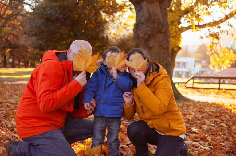 a picture of a family of three in bright jackets holding autumn leaves in front of their faces